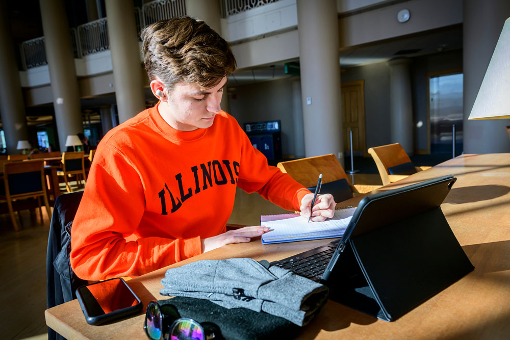 student wearing bright orange Illinois sweatshirt studies with laptop in a library