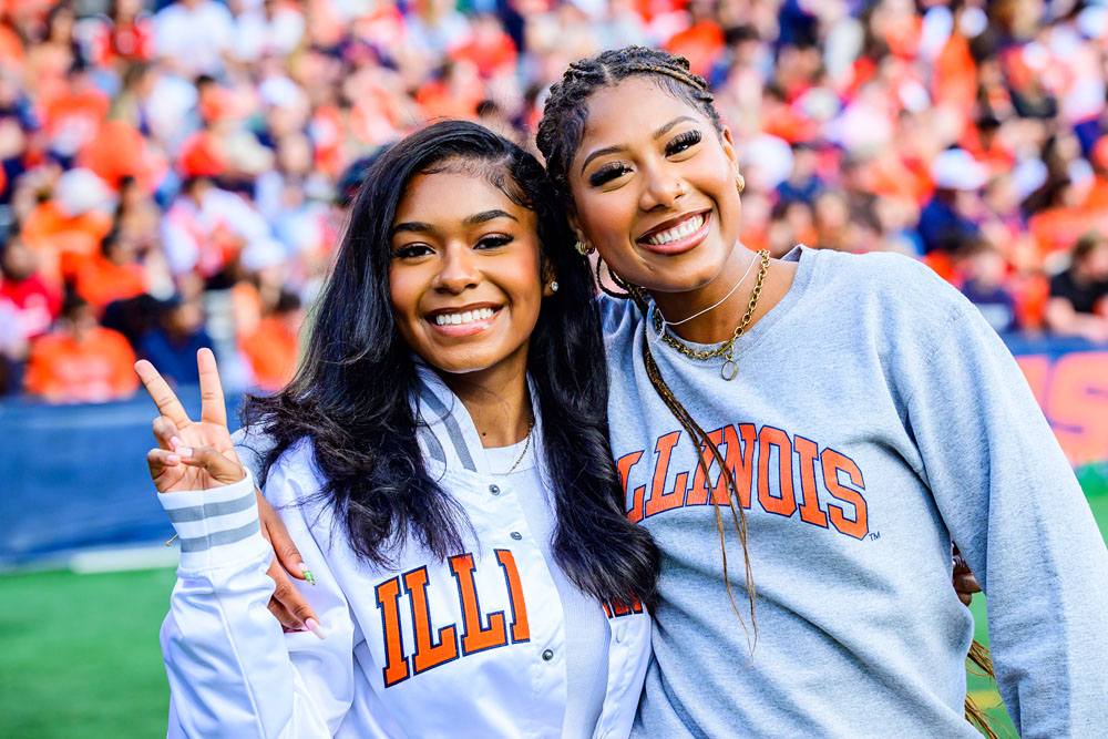 two smiling friends hug and pose in Illinois apparel on the field for gameday