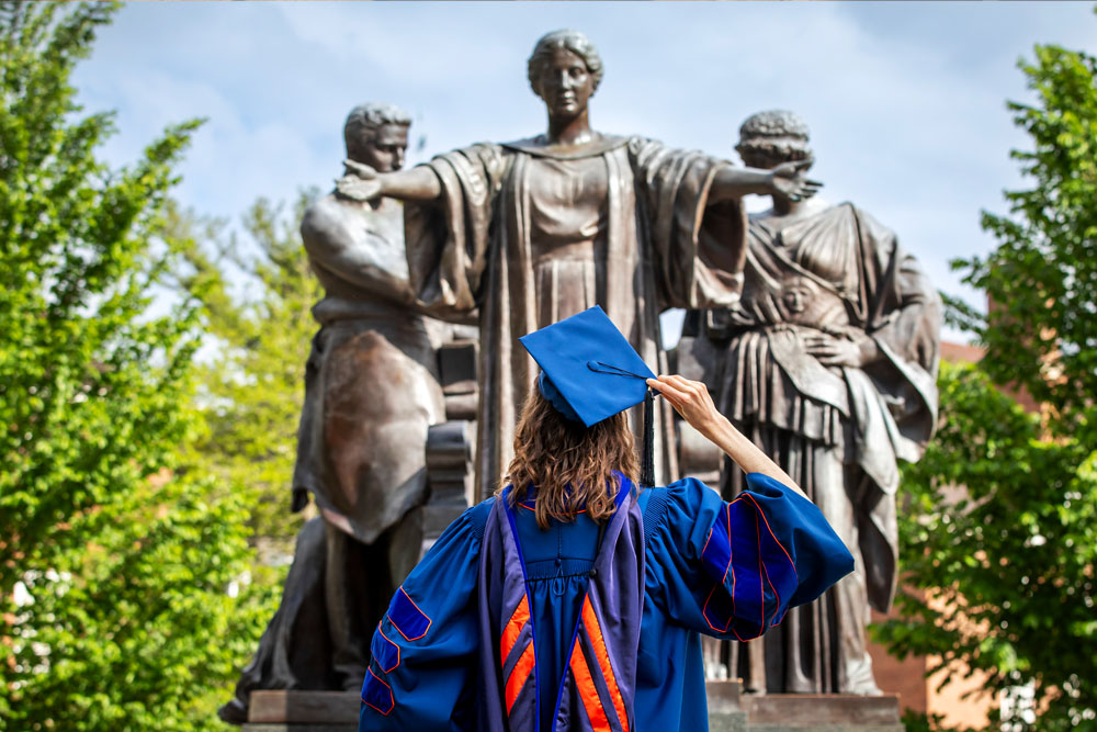 recent graduate in full orange and blue regalia faces Alma Mater statue and moves to remove their cap