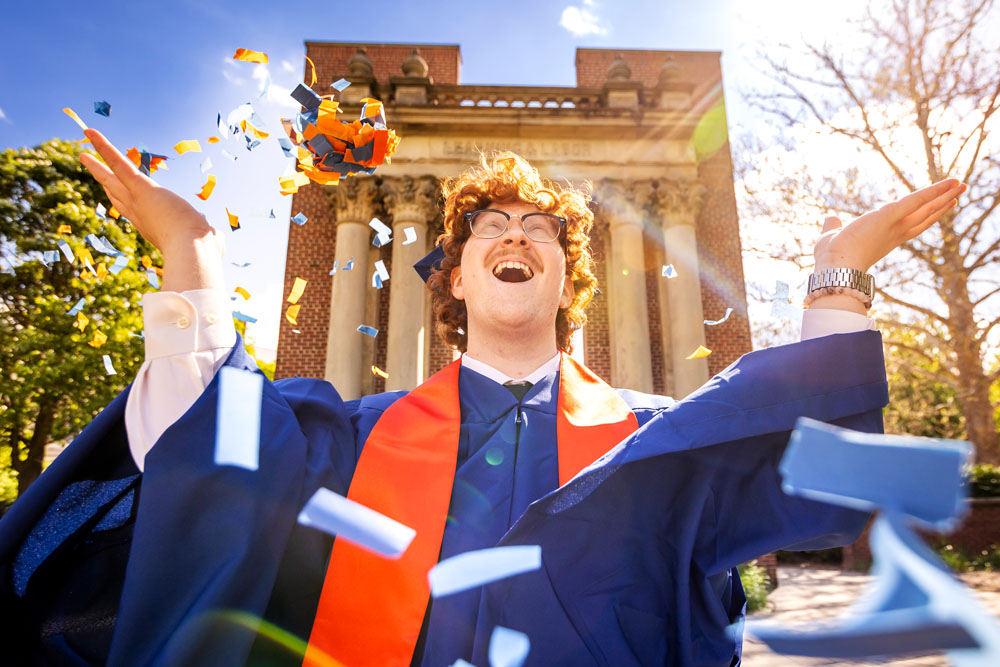 graduating Illini poses in front of Hallene Gateway with thrown orange and blue confetti