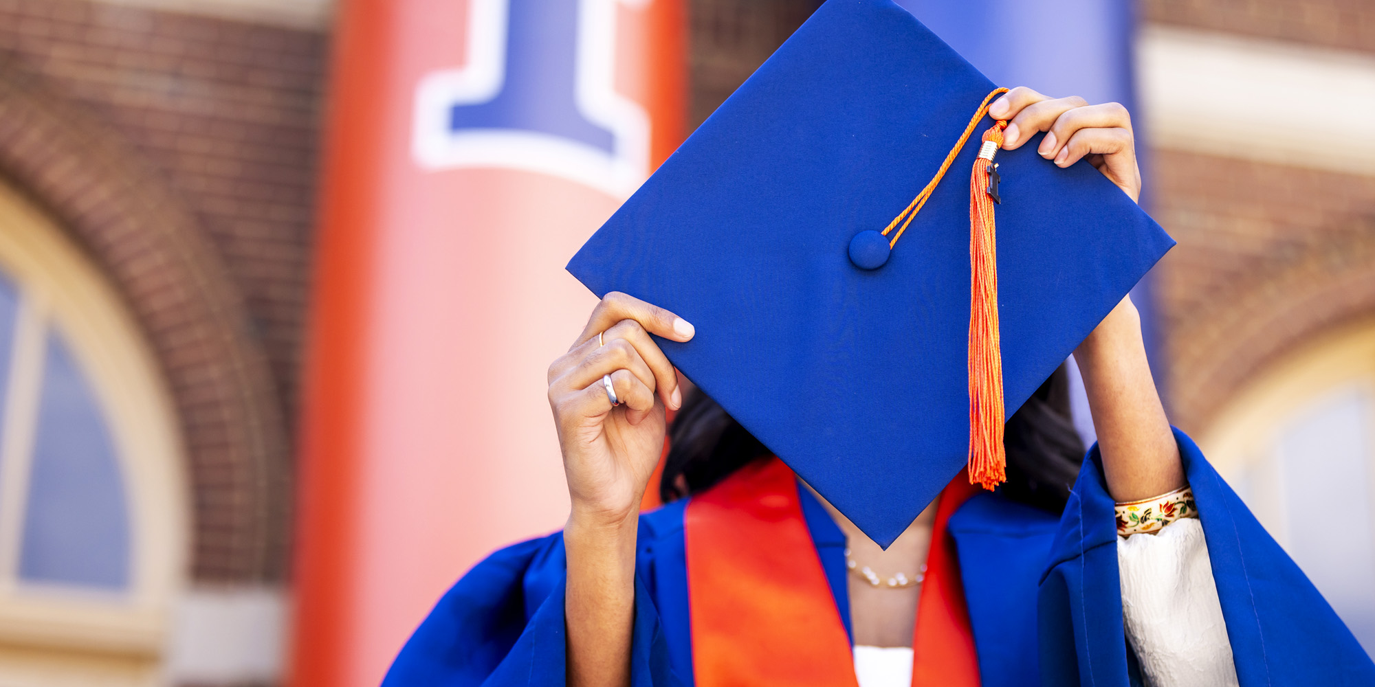 graduate in orange and blue regalia on the steps of Foellinger Auditorium hiding their face behind cap and tassle
