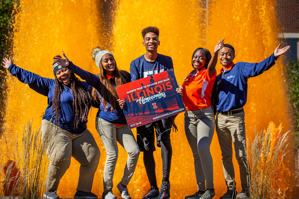 students in orange and blue pose in front of the Alice Campbell Alumni Center fountain, dyed orange for Homecoming