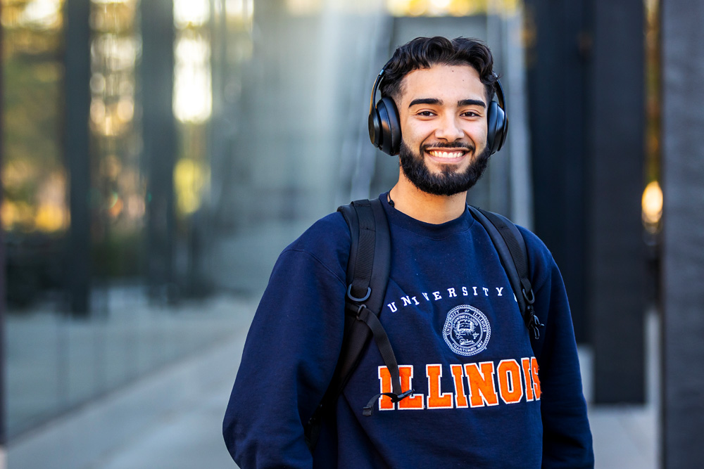 student wearing blue Illinois sweatshirt and headphones smiles outside the Campus Instructional Facility off the Grainger College of Engineering's Bardeen Quad