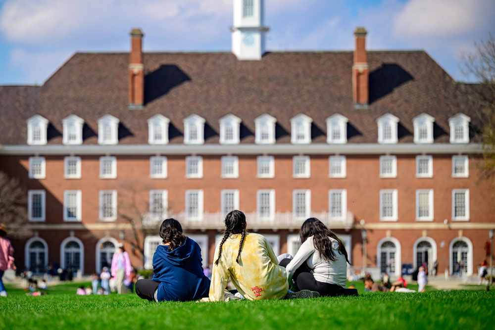 three students sit on the grass on the Quad with the Illini Union towering in the background