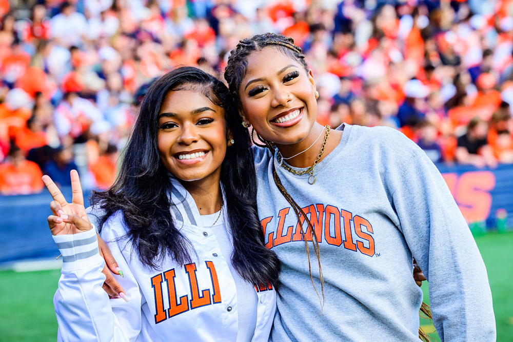 friends wearing Illinois sweatshirts pose on-field during an Illini football game at Gies Memorial Stadium